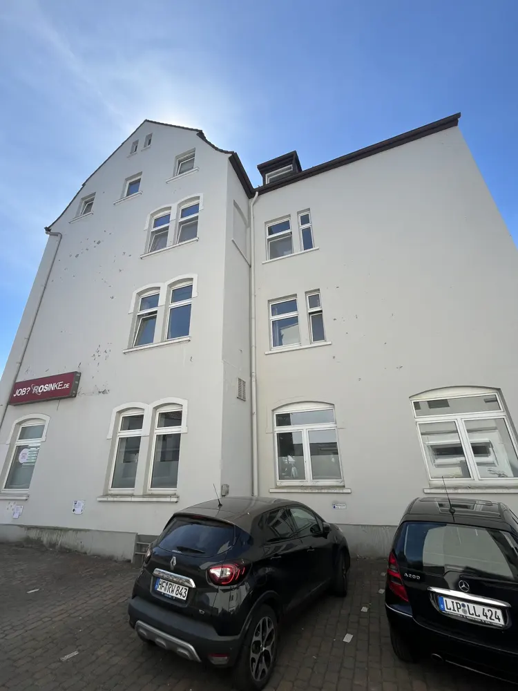 Exterior view of a tall, light gray building with many windows under a blue sky. Two black cars are parked in front. A sign reads "JOB? ROSINKE.de".
