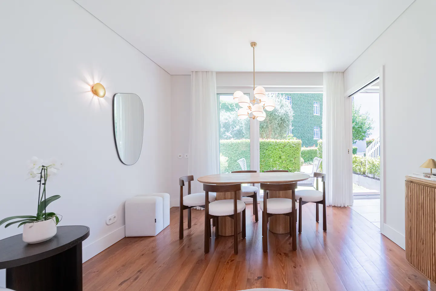 Bright dining room with wood floors, white walls, and a view of a green garden through a large window.