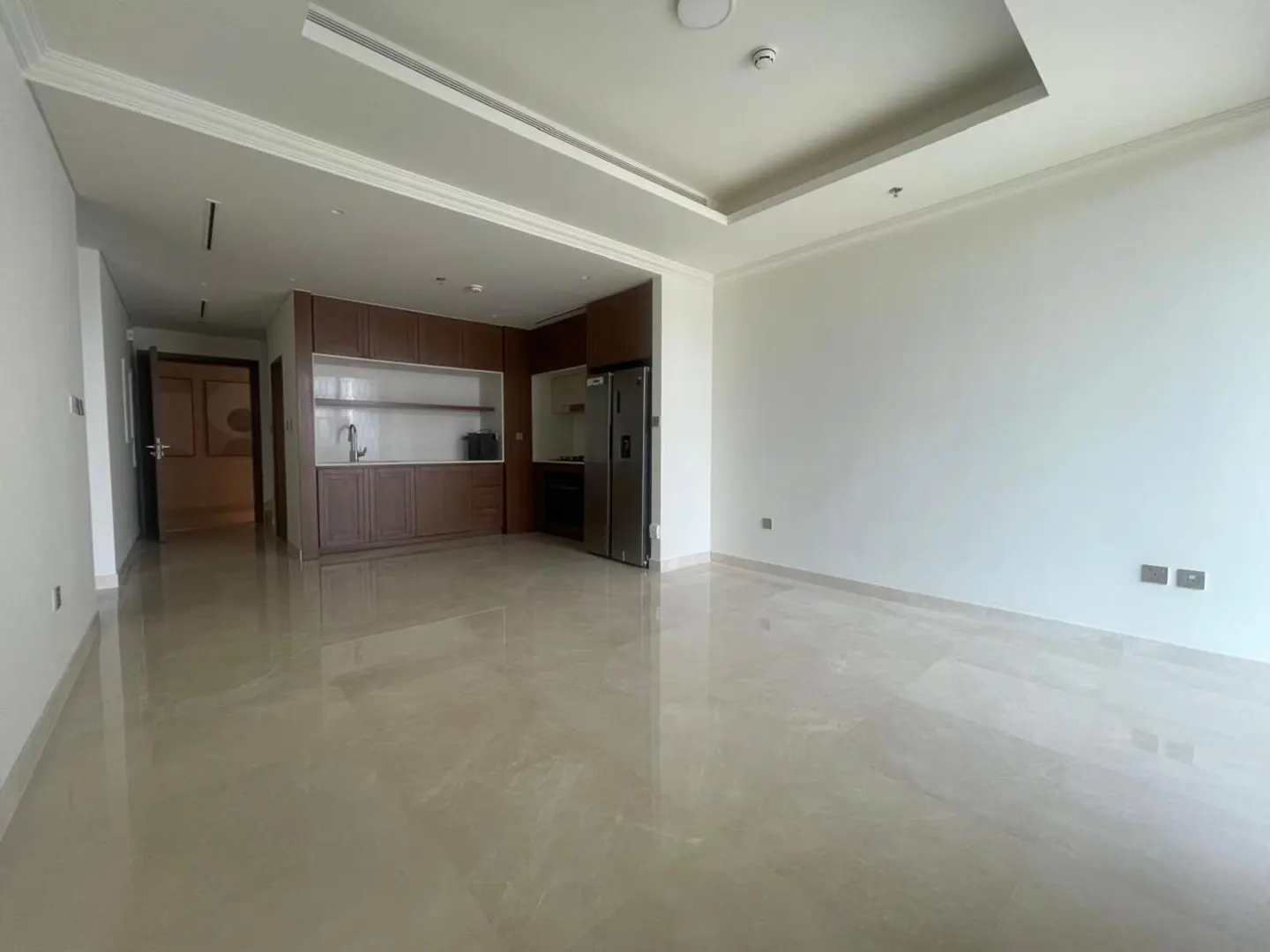 Modern open-plan living area with white walls, glossy tile floors, and a kitchen featuring dark wood cabinets and a fridge.