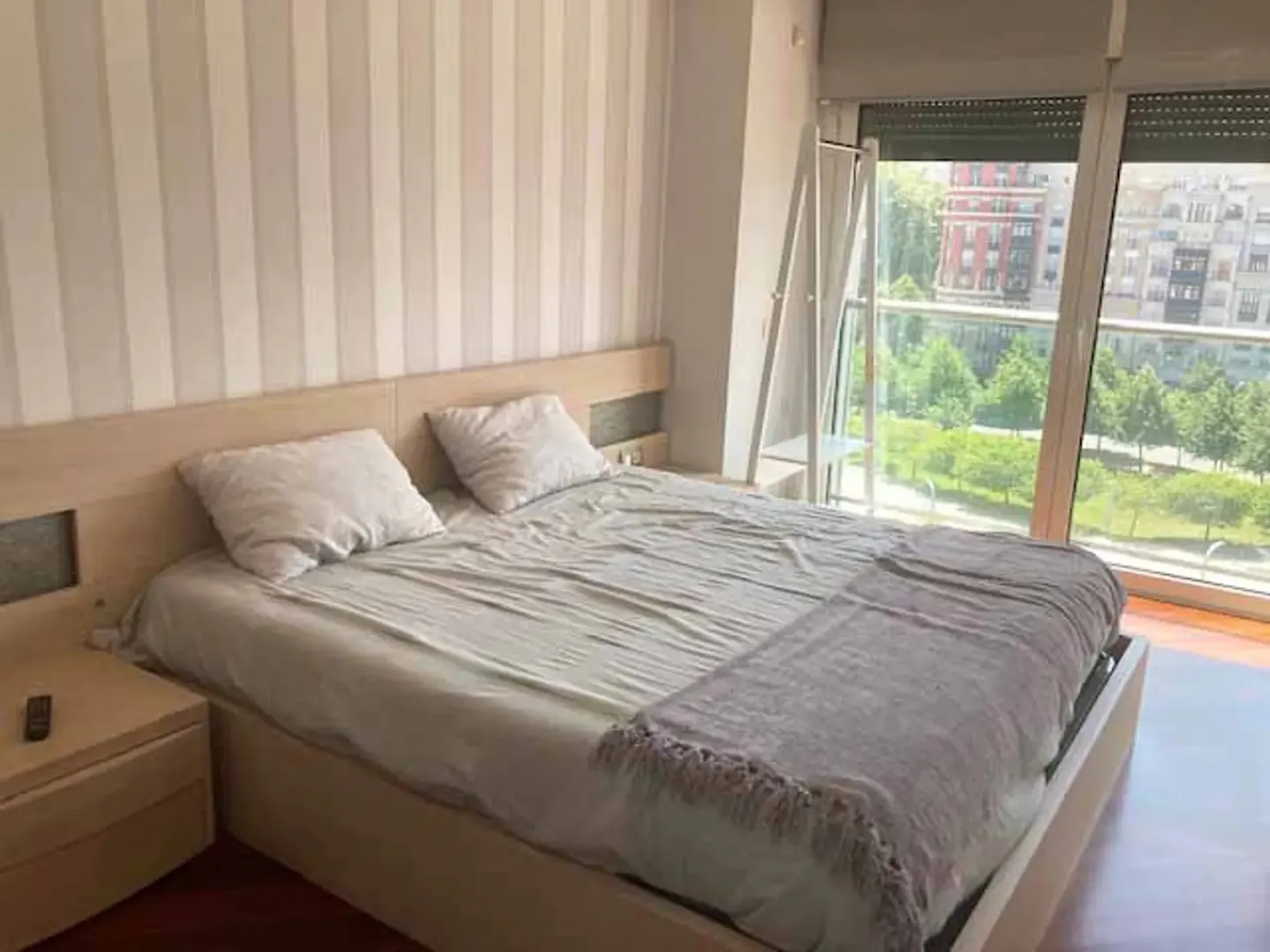 Bedroom with a light wood bed, striped wallpaper, and a large window overlooking a green park and city buildings.