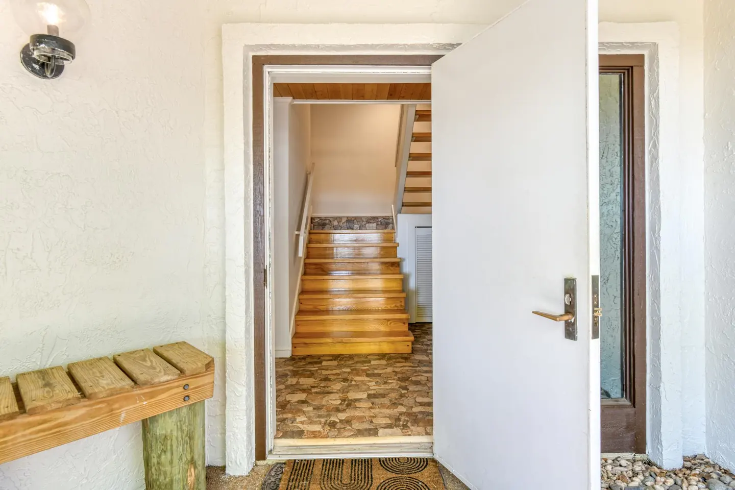 Open white door reveals a staircase with wooden steps and patterned flooring. A wooden bench sits to the left of the doorway.