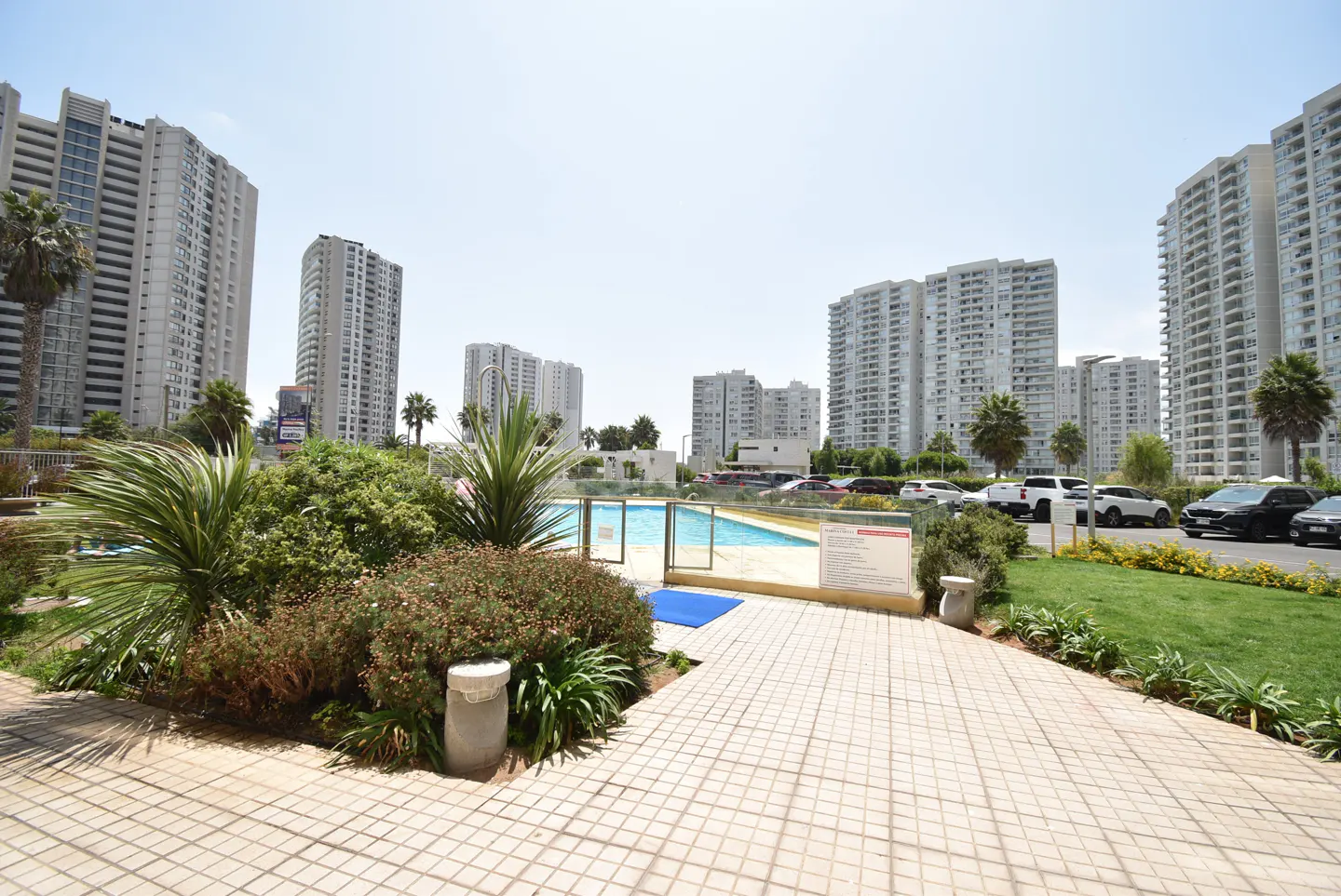 Outdoor pool area with tiled patio, green plants, and tall apartment buildings in the background on a sunny day.