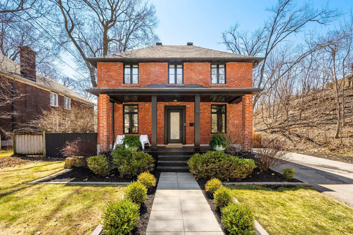 Two-story red brick house with a black porch and door. A stone walkway leads to the house, surrounded by green bushes and grass.