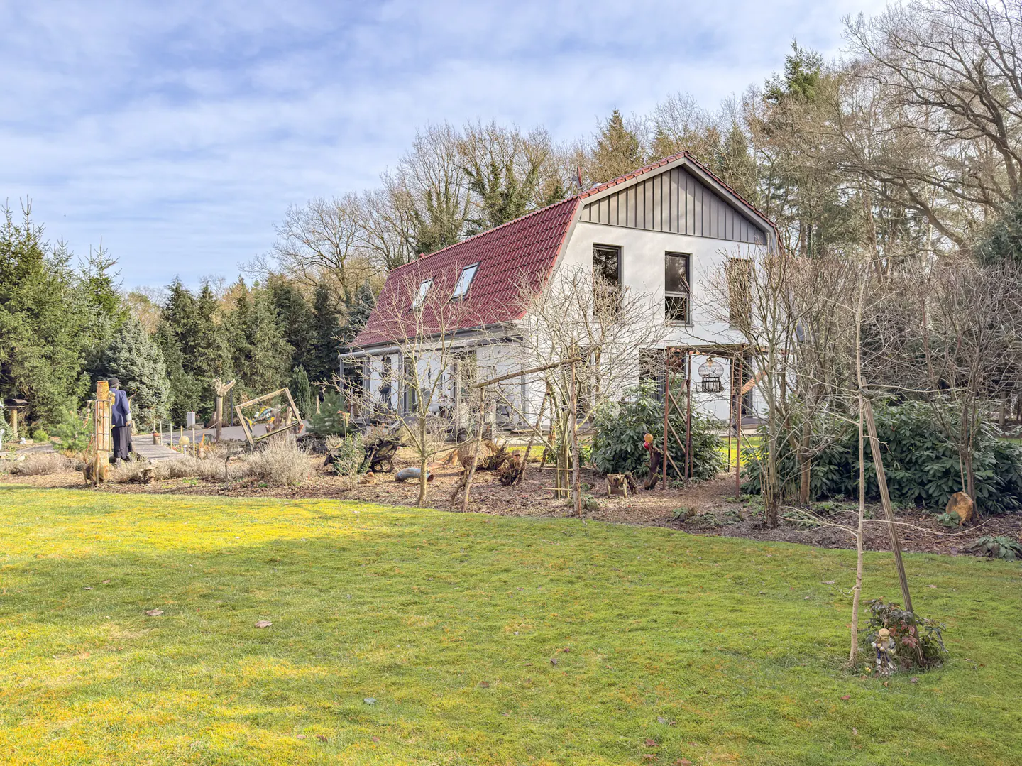 Two-story white house with a red roof, surrounded by trees and a green lawn. A person walks along a path in the yard.