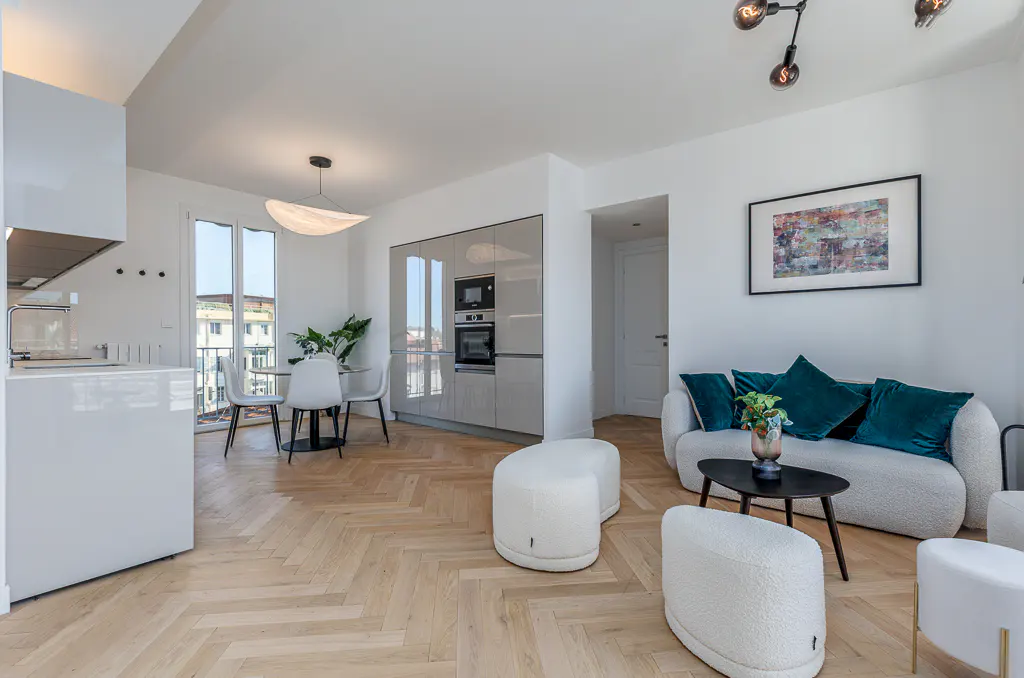 Bright, modern apartment interior with white walls, herringbone wood floors, and stylish furniture. Kitchen, dining, and living areas are visible.