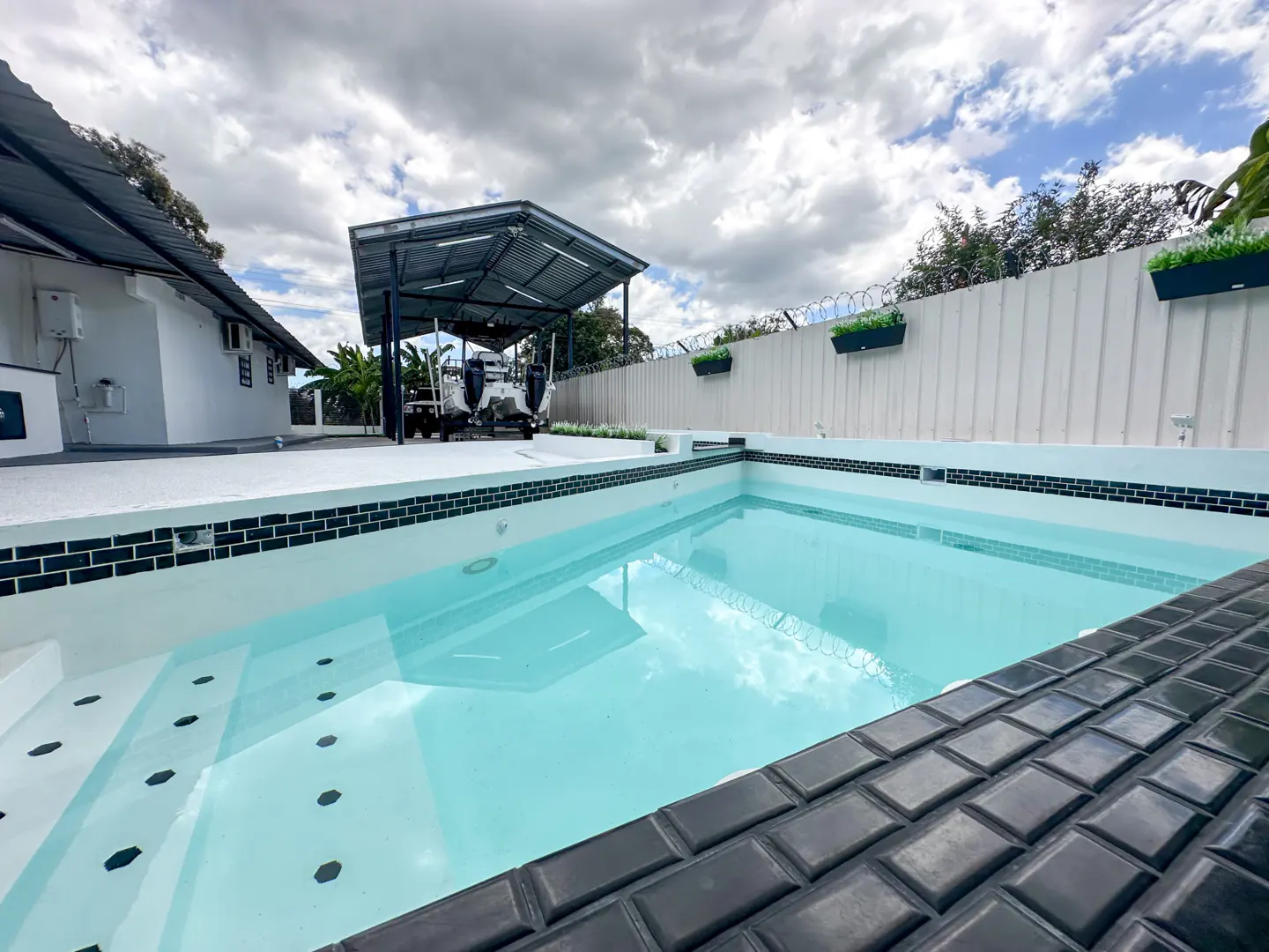 A backyard pool with black tile trim and clear blue water, next to a white house and boat shelter.