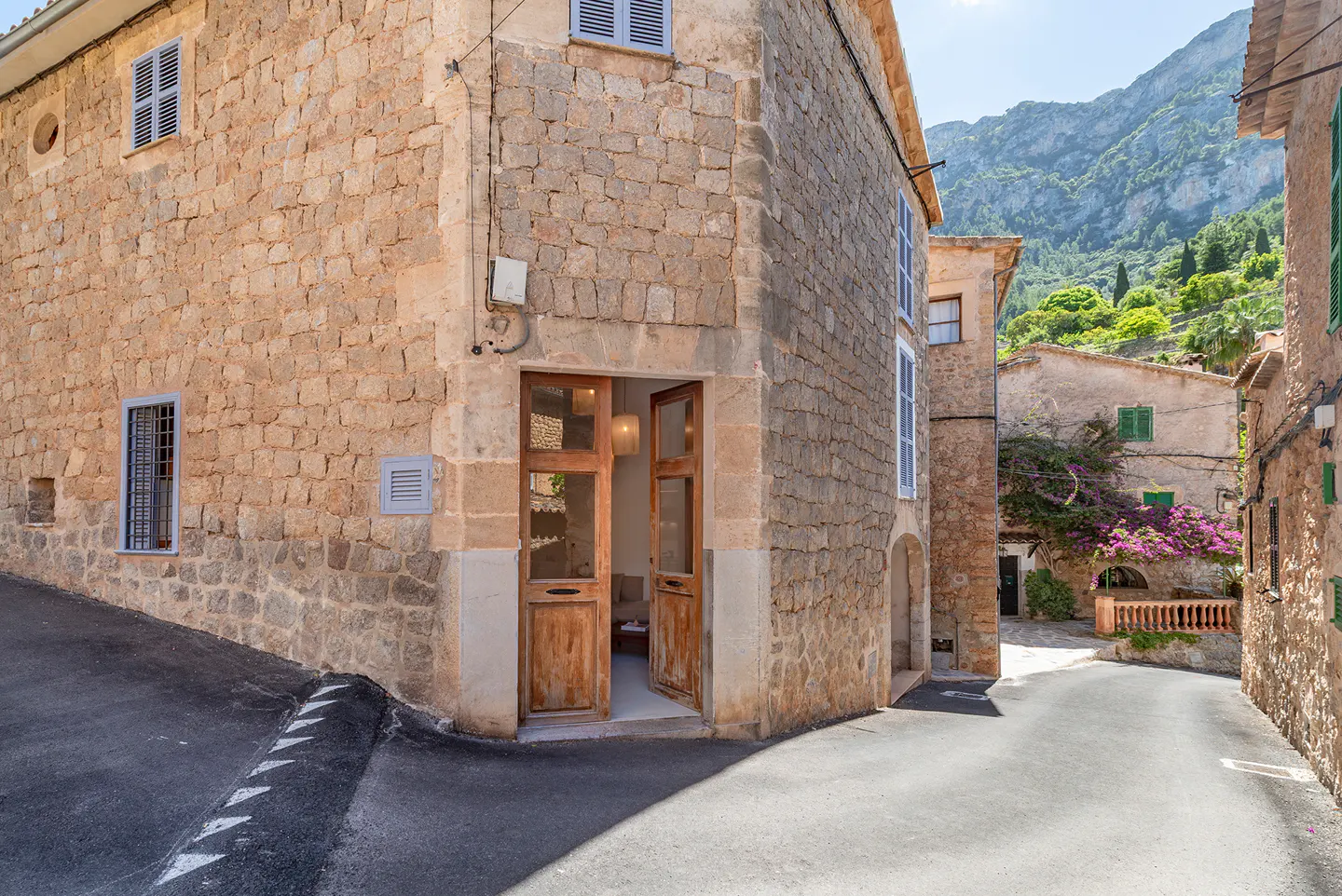 Exterior view of a stone building with an open wooden door, revealing a glimpse of the interior. Mountains are visible in the background.