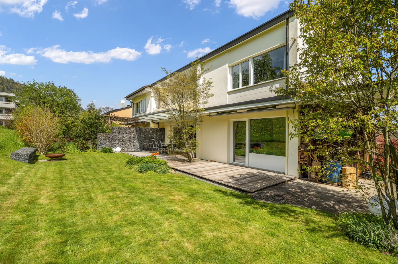 Two-story house with a green lawn, stone wall, and wooden deck on a sunny day.