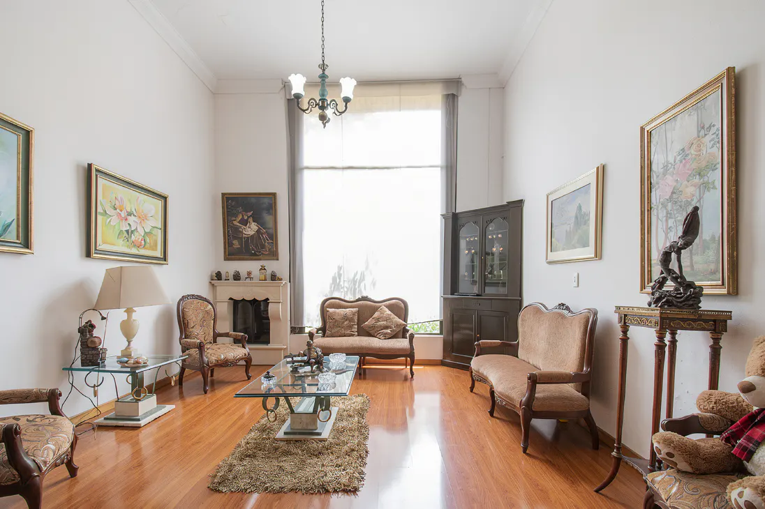 Bright living room with hardwood floors, white walls, and tall windows. Antique furniture, paintings, and a fireplace add character.