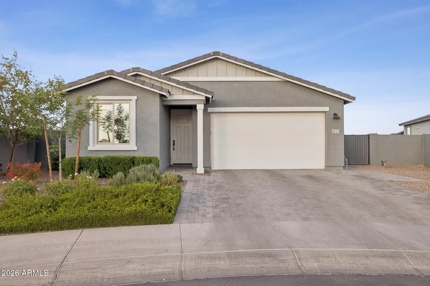 A gray single-story house with a white garage door and front door, green bushes, and a blue sky.