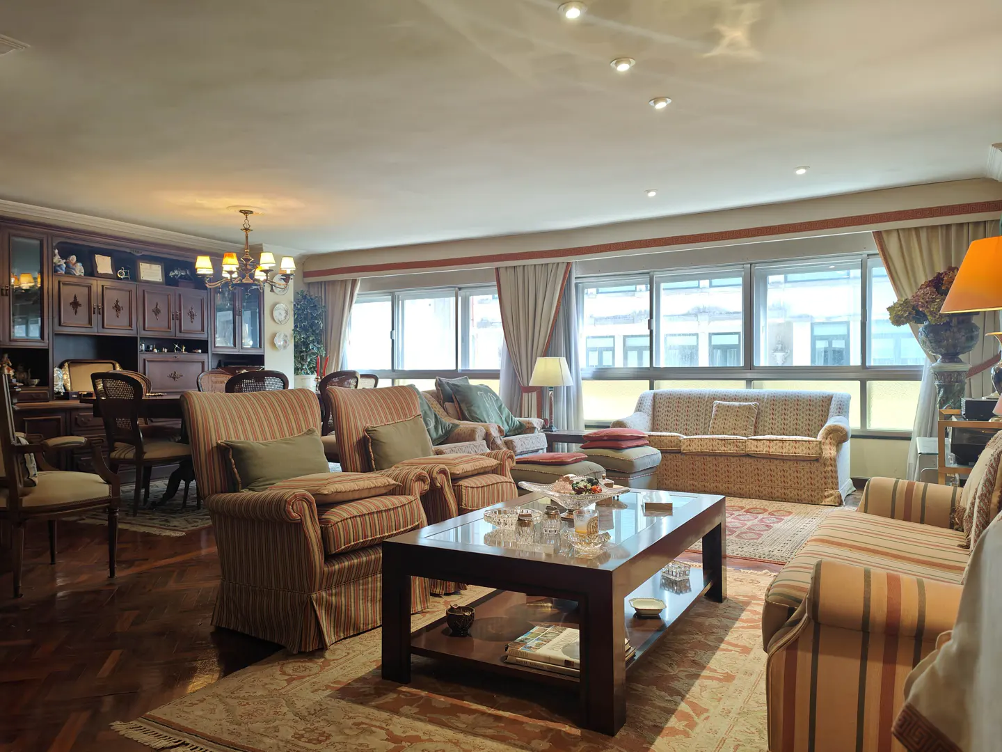 Spacious living room with striped armchairs, a dark wood coffee table, and large windows with beige curtains.