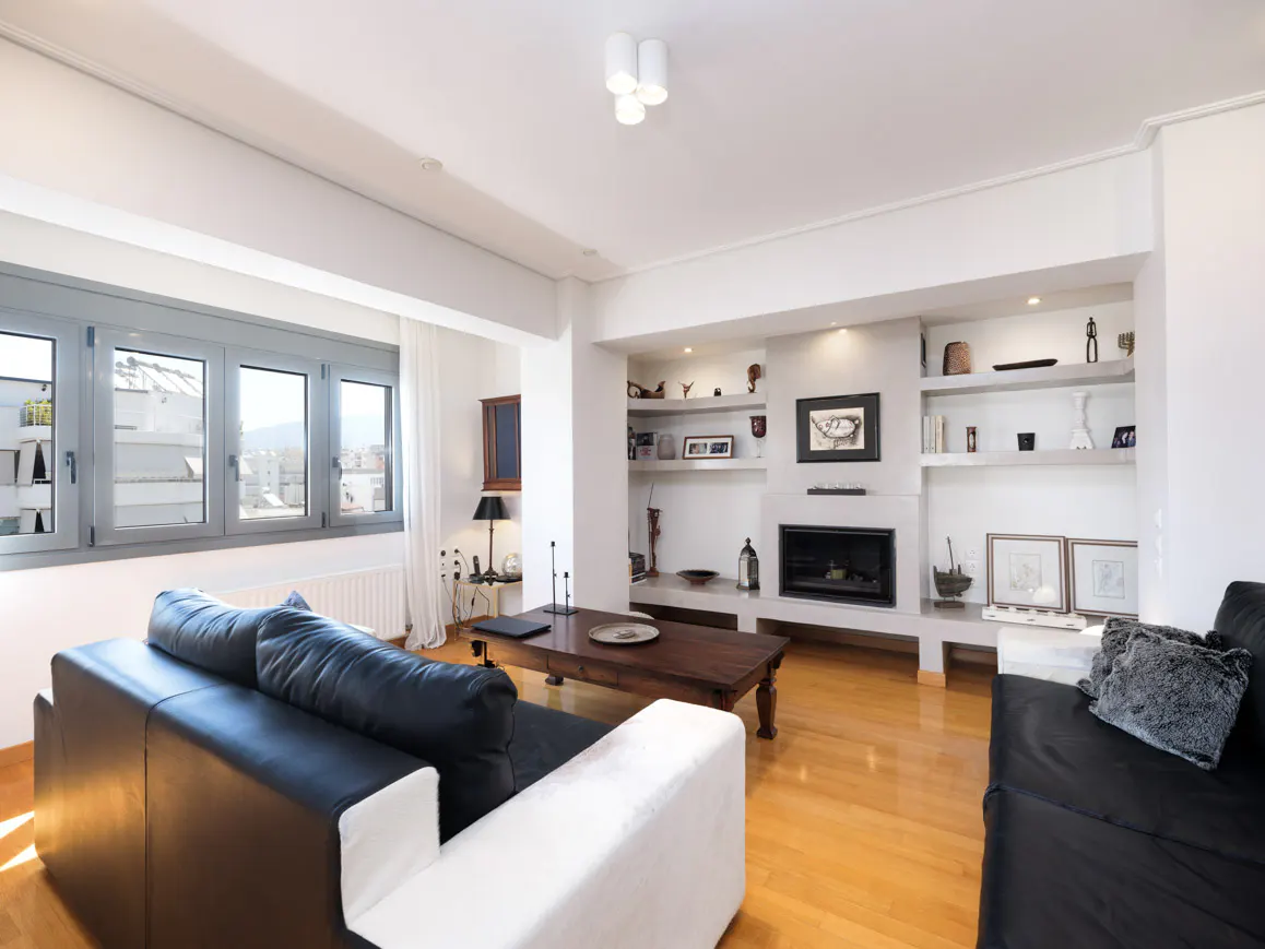 Bright living room with hardwood floors, black leather sofas, and a dark wood coffee table. A fireplace is surrounded by built-in shelves.