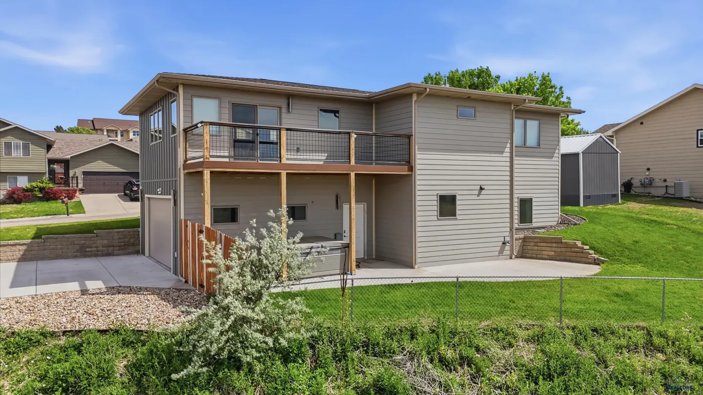 Two-story gray house with a wooden deck, a ground-level patio, a hot tub, and a fenced backyard on a sunny day.