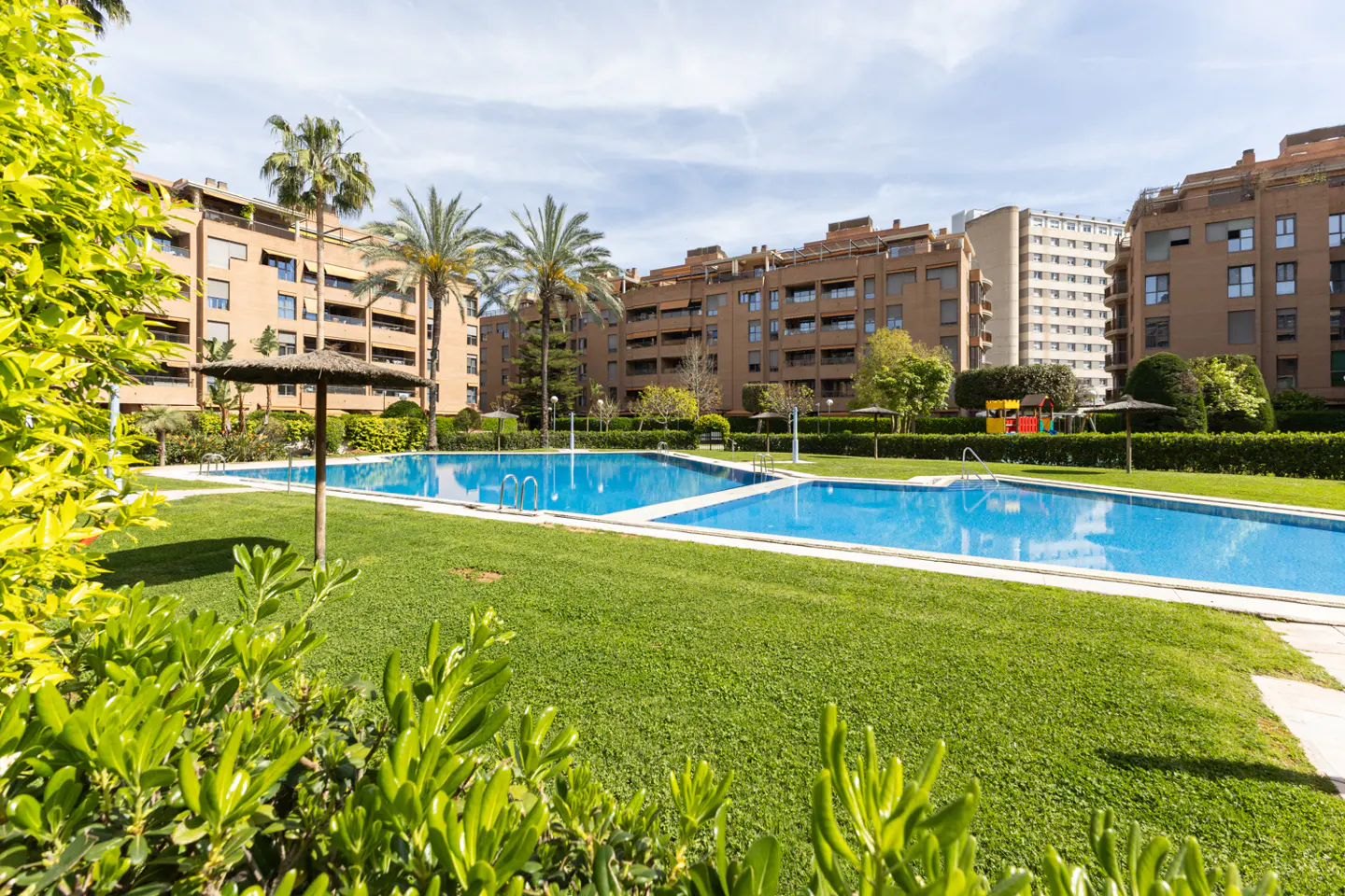Outdoor pool area with blue water, green grass, palm trees, and apartment buildings in the background on a sunny day.