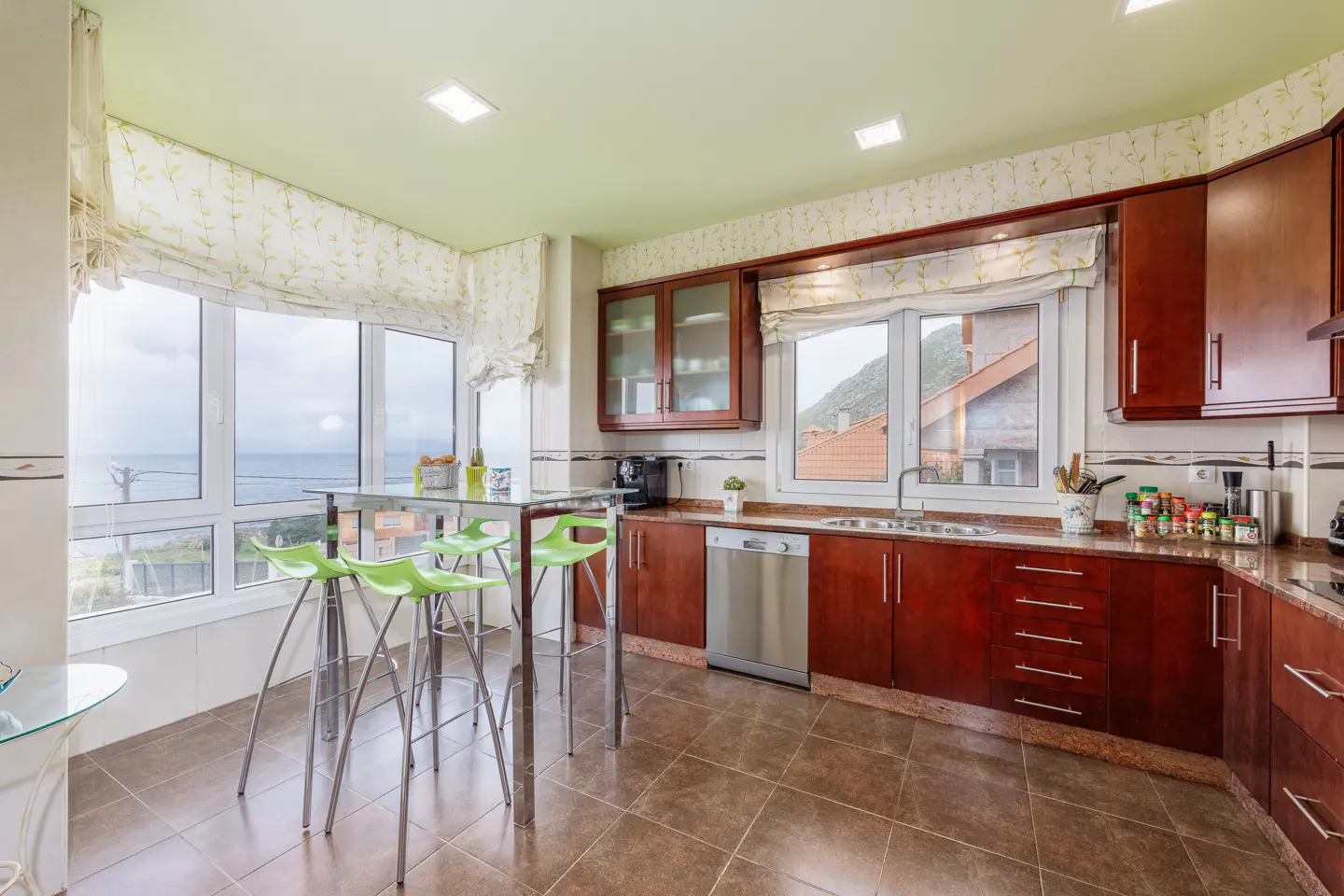Bright kitchen with cherry cabinets, brown tile floor, and a glass table with four green stools. Large windows offer a view of the ocean.