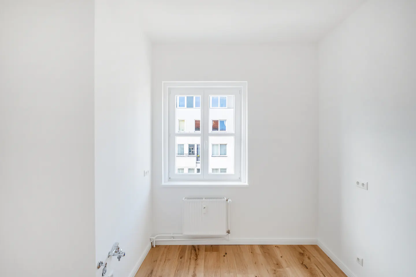 Bright, empty room with light wood floors and white walls. A white window looks out onto a building. A white radiator sits below the window.