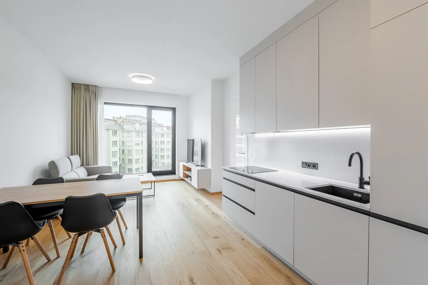 Modern open-plan apartment with a white kitchen, dining area with black chairs, and a living space with city views.