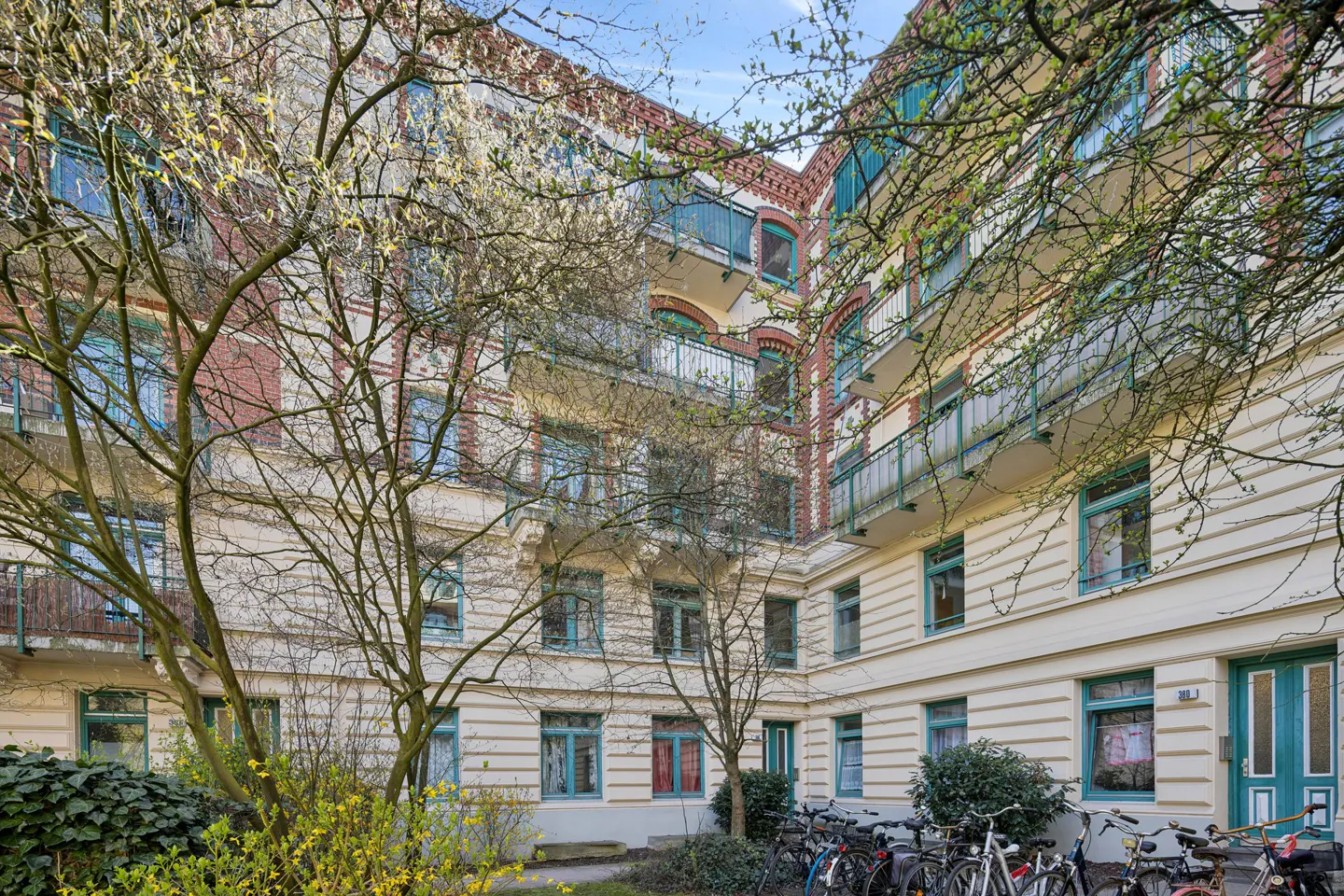 Exterior view of a cream-colored apartment building with turquoise window frames and balconies, framed by trees and parked bicycles.