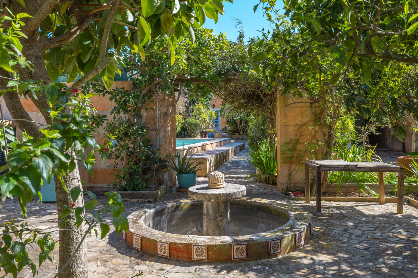 Stone courtyard with a tiled fountain, lush greenery, and a glimpse of a pool in the background. A wooden table sits to the right.
