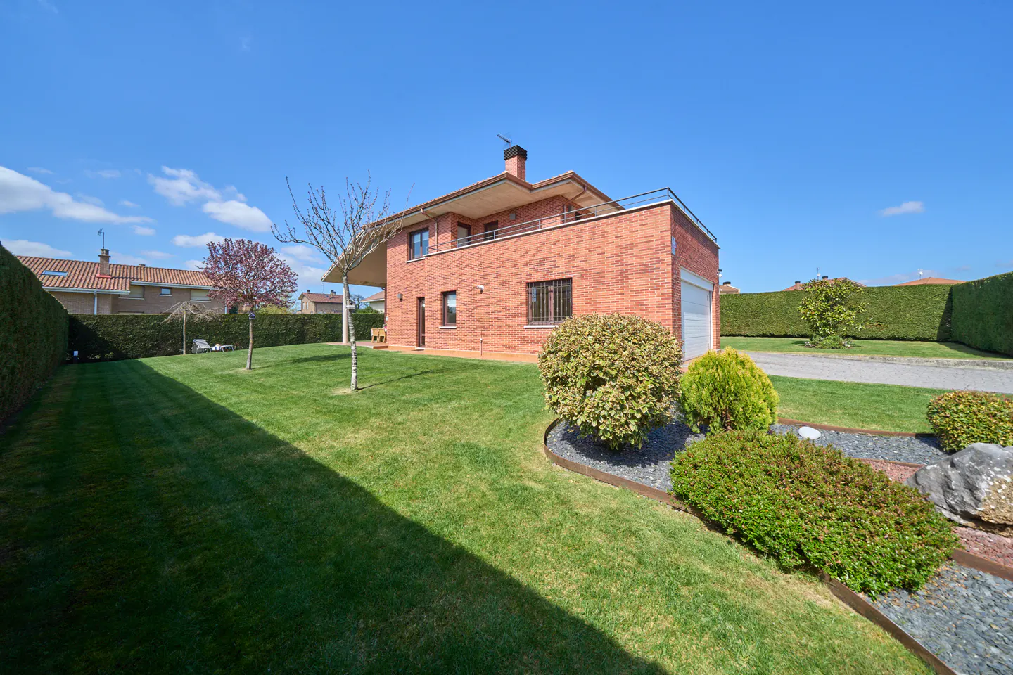 Two-story brick house with a balcony, green lawn, and trimmed hedges under a blue sky.