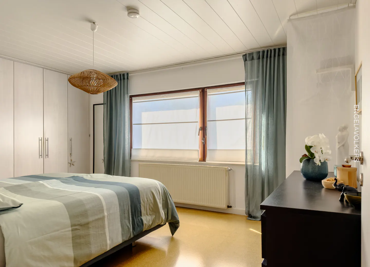 Bright bedroom with a striped bed, white built-in closets, a woven pendant light, and blue curtains over a large window.