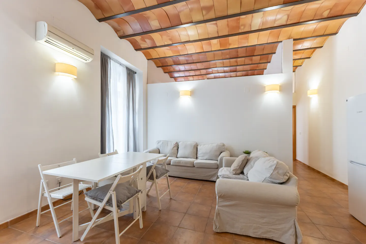 Bright living space with white walls, terracotta tile floor, and a unique terracotta-tiled ceiling. A white table and sofas furnish the room.