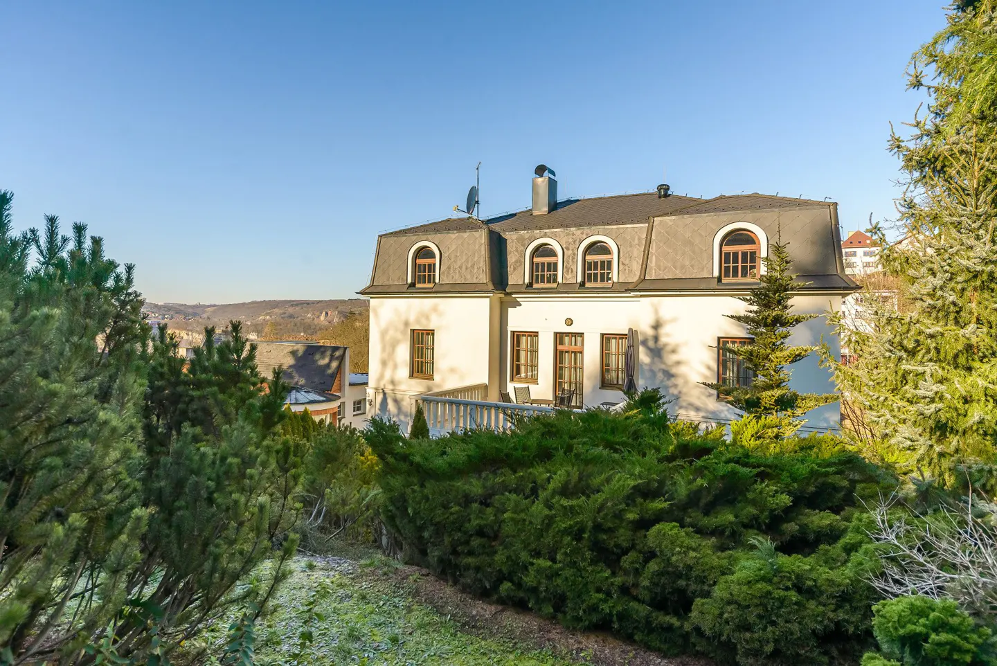 Exterior view of a white two-story house with a gray roof and arched windows, surrounded by green trees and bushes.