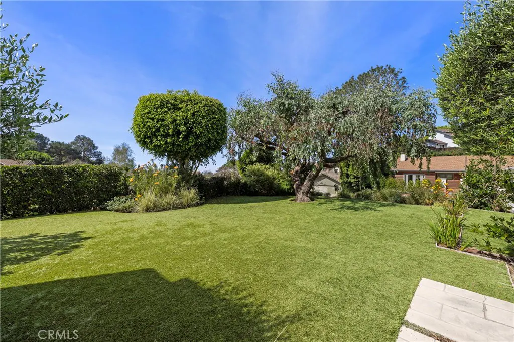 A lush green lawn with trees and hedges under a clear blue sky. A house is visible in the background.