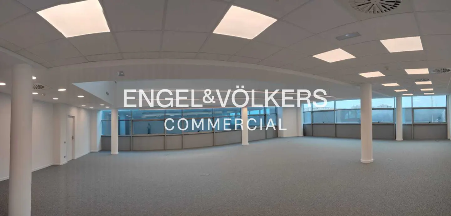 Empty commercial space with gray carpet, white walls, and large windows. Engel & Völkers Commercial logo overlay.