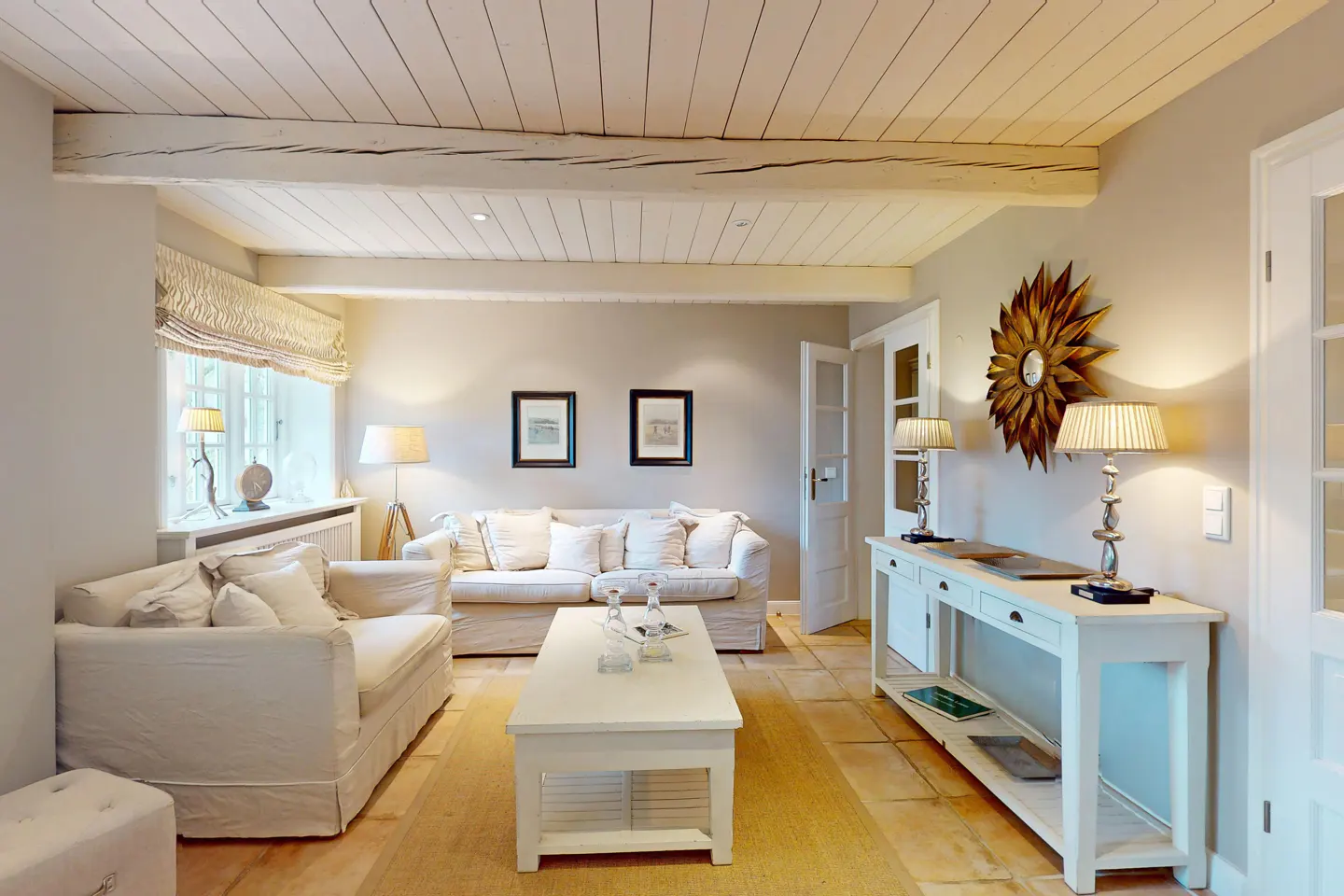 Bright living room with white sofas, coffee table, and console table. Wooden ceiling beams and a sunburst mirror add character.