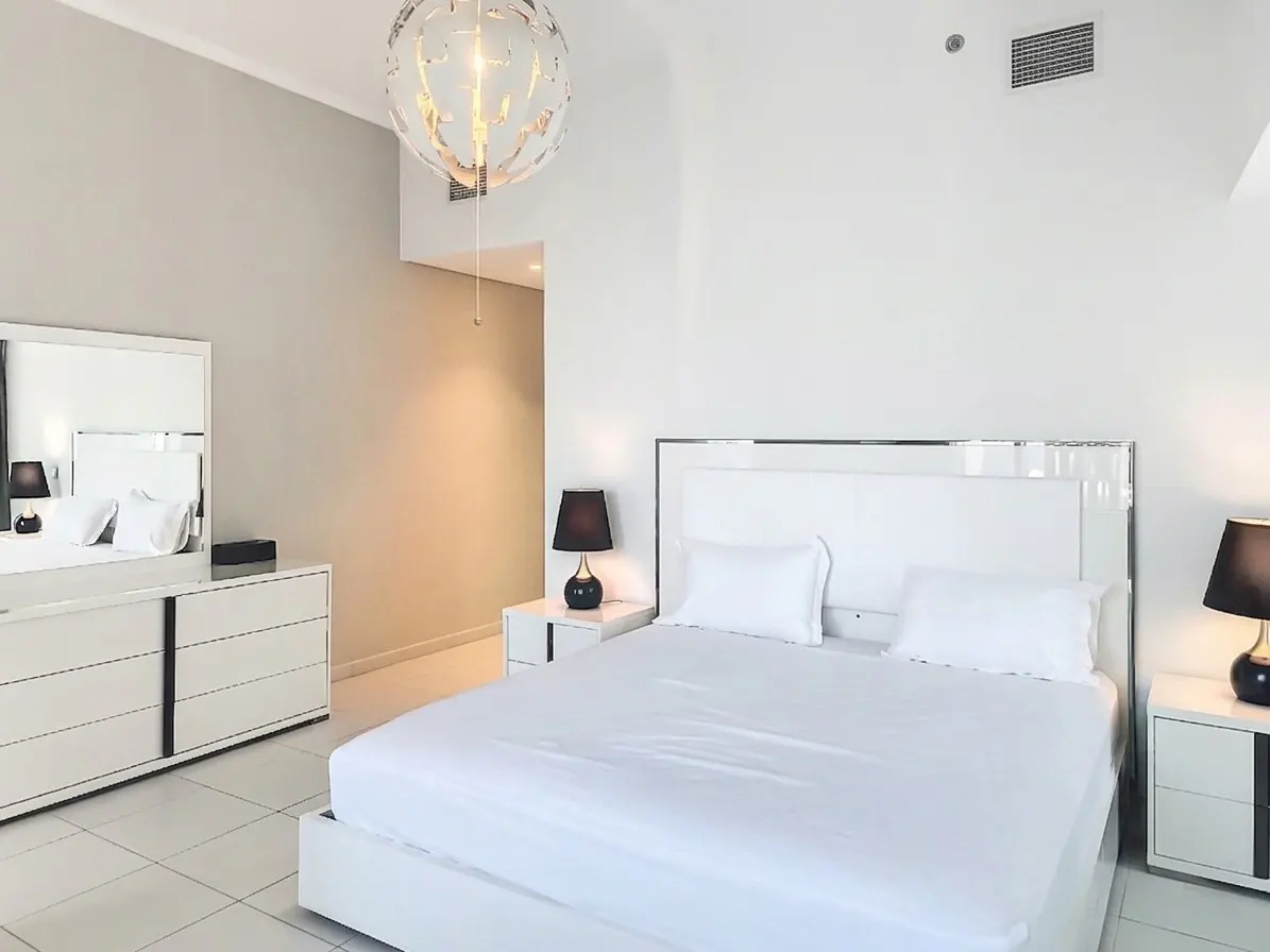Bright bedroom with white bed, dresser, and nightstands. A modern chandelier hangs from the ceiling.