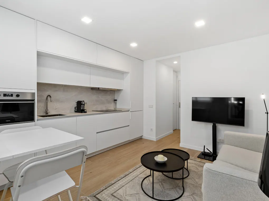 Bright, modern apartment with white kitchen, table, black coffee tables, and a TV. A patterned rug adds texture to the light wood floor.