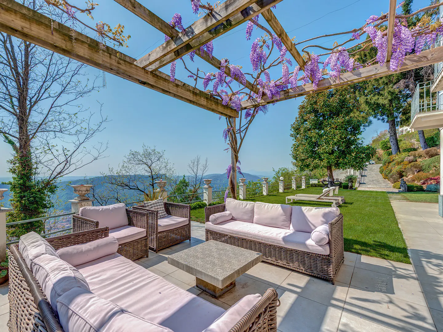 Outdoor patio with wicker furniture and light purple cushions under a pergola with wisteria. Rolling hills in the background.