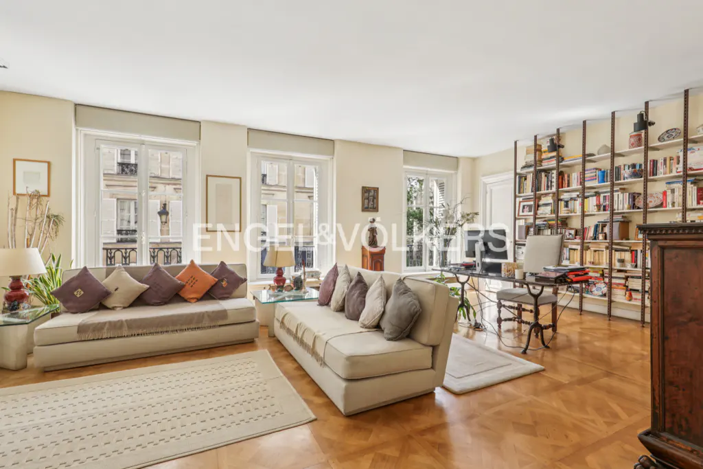 Bright living room with hardwood floors, two sofas with throw pillows, a desk, and a large bookshelf. Windows overlook a city street.