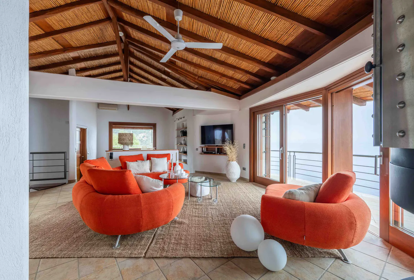 Bright living room with orange sofas, white walls, and a straw ceiling with a fan. Sliding glass doors lead to a balcony with an ocean view.