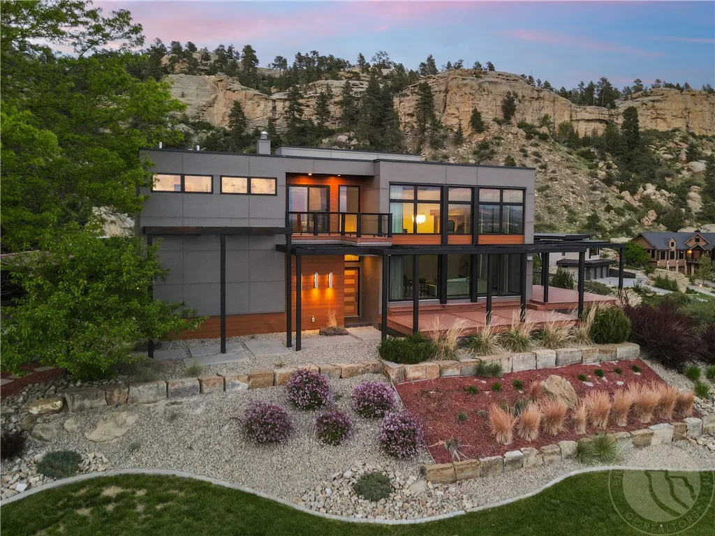 Modern gray house with black trim and large windows, set against a mountain backdrop at dusk. Landscaped yard with flowers and rocks.