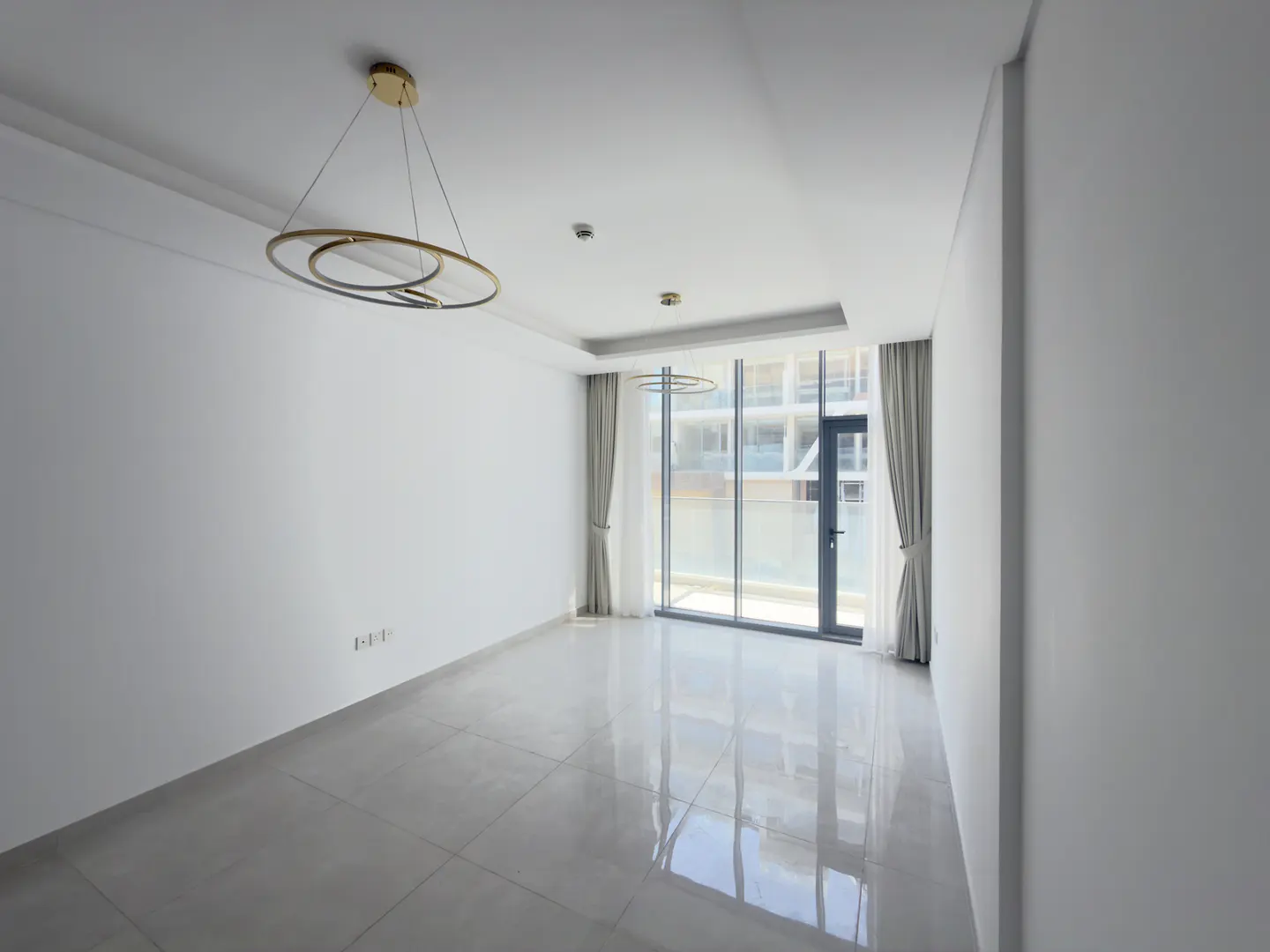 Minimalist empty room with white walls, glossy tile floors, a modern gold ring chandelier, and floor-to-ceiling windows.