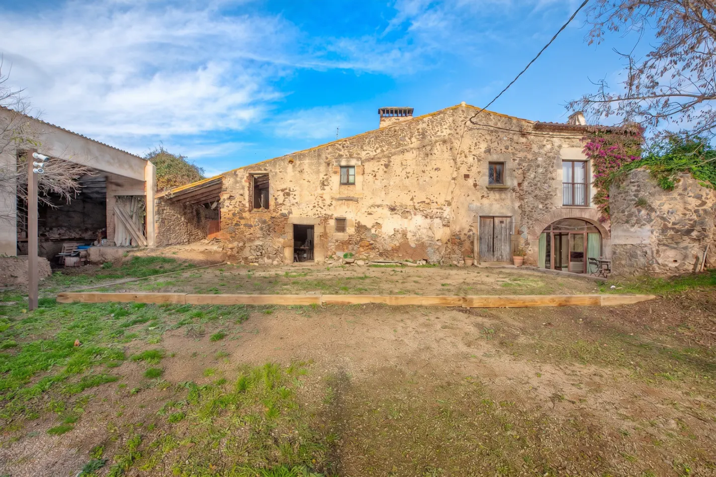 Exterior view of a stone farmhouse with a barn under a blue sky. The house has a chimney and multiple windows.