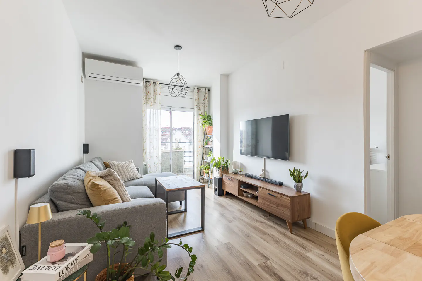 Bright living room with gray sectional sofa, wood floors, TV, and balcony. Plants add a touch of nature to the modern space.