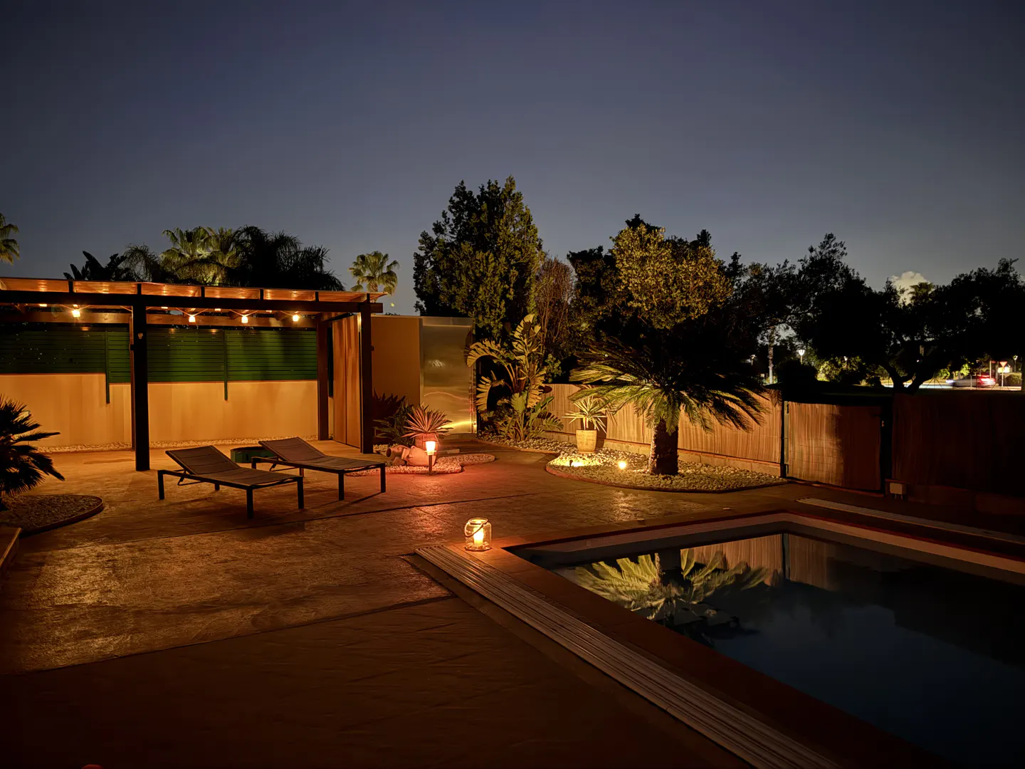 Night view of a backyard with a pool, lounge chairs, and palm trees, illuminated by warm lights.