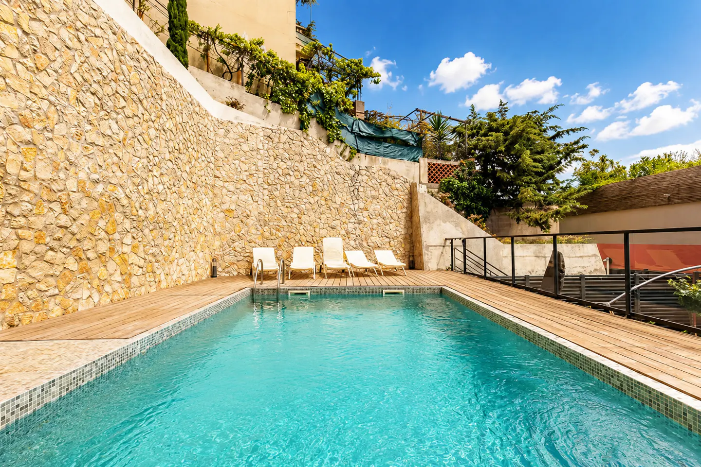 Outdoor pool with turquoise water, surrounded by a wooden deck and lounge chairs, set against a stone wall and blue sky.