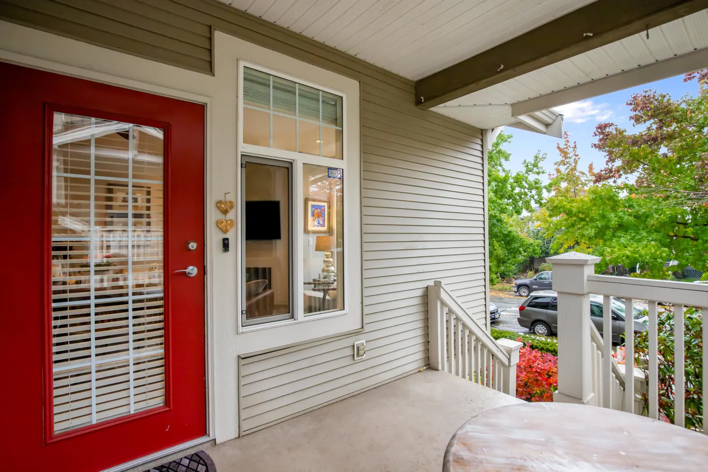 Exterior view of a home's porch with a red door, white railing, and a glimpse of green trees in the background.