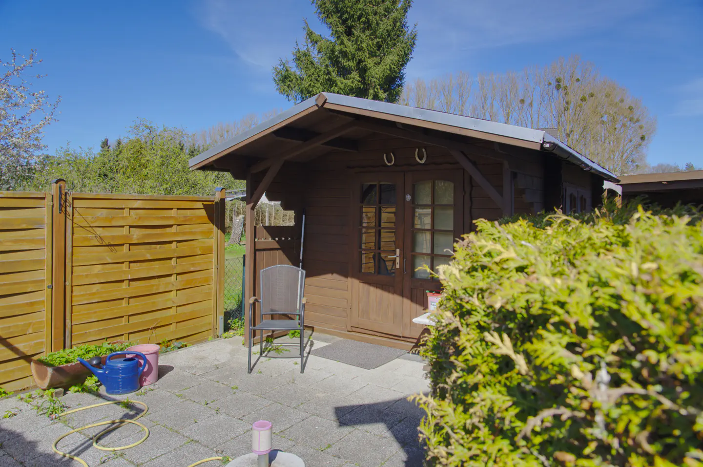 A brown wooden shed with a chair on a stone patio, a wooden fence, and green bushes.