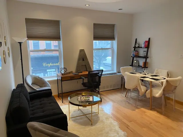 Living room with hardwood floors, black sofa, desk by window, and dining table with four white chairs.