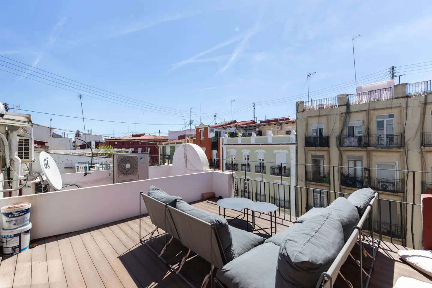 Rooftop patio with gray lounge seating and a small table. City buildings are visible in the background under a blue sky.