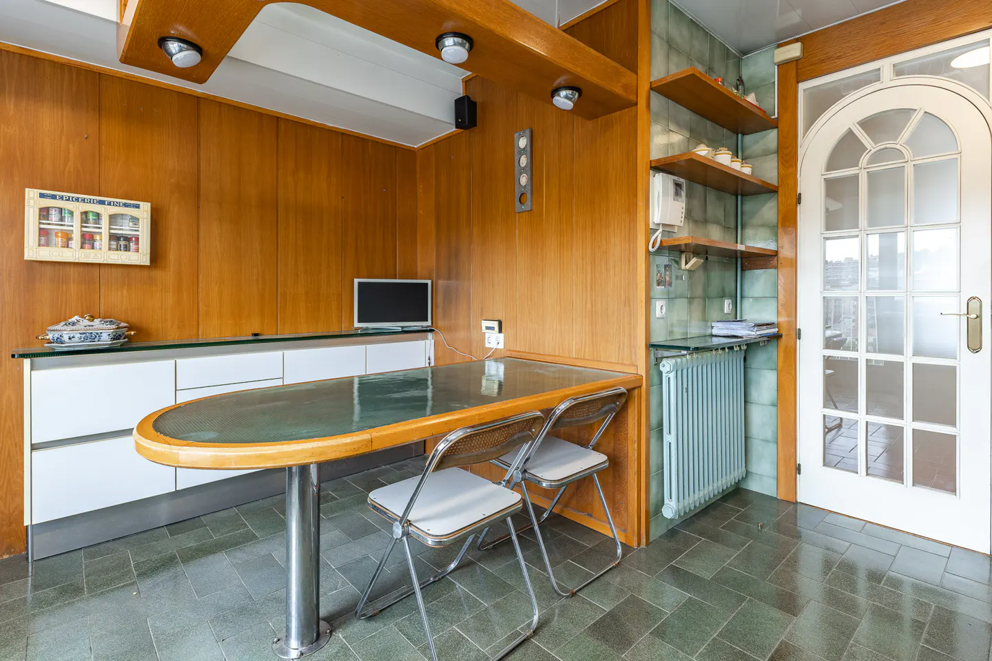 A kitchen with wood paneling, a glass-topped table, and two chairs. A white door with glass panes is on the right.