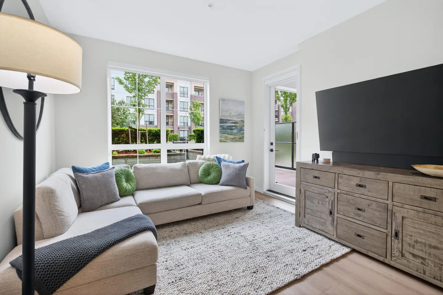 Modern living room with a beige sectional sofa, grey rug, rustic wooden media console, large TV, and a floor lamp.