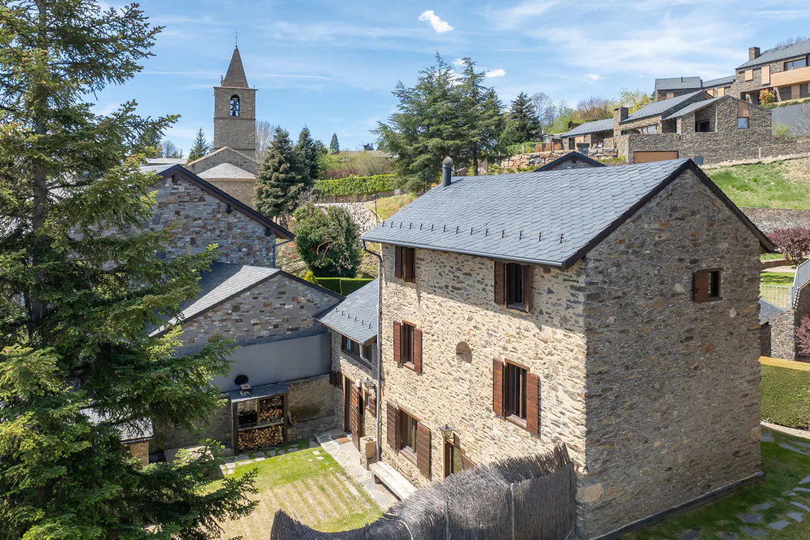 Stone house with brown shutters and a gray roof, with a church steeple in the background on a sunny day.