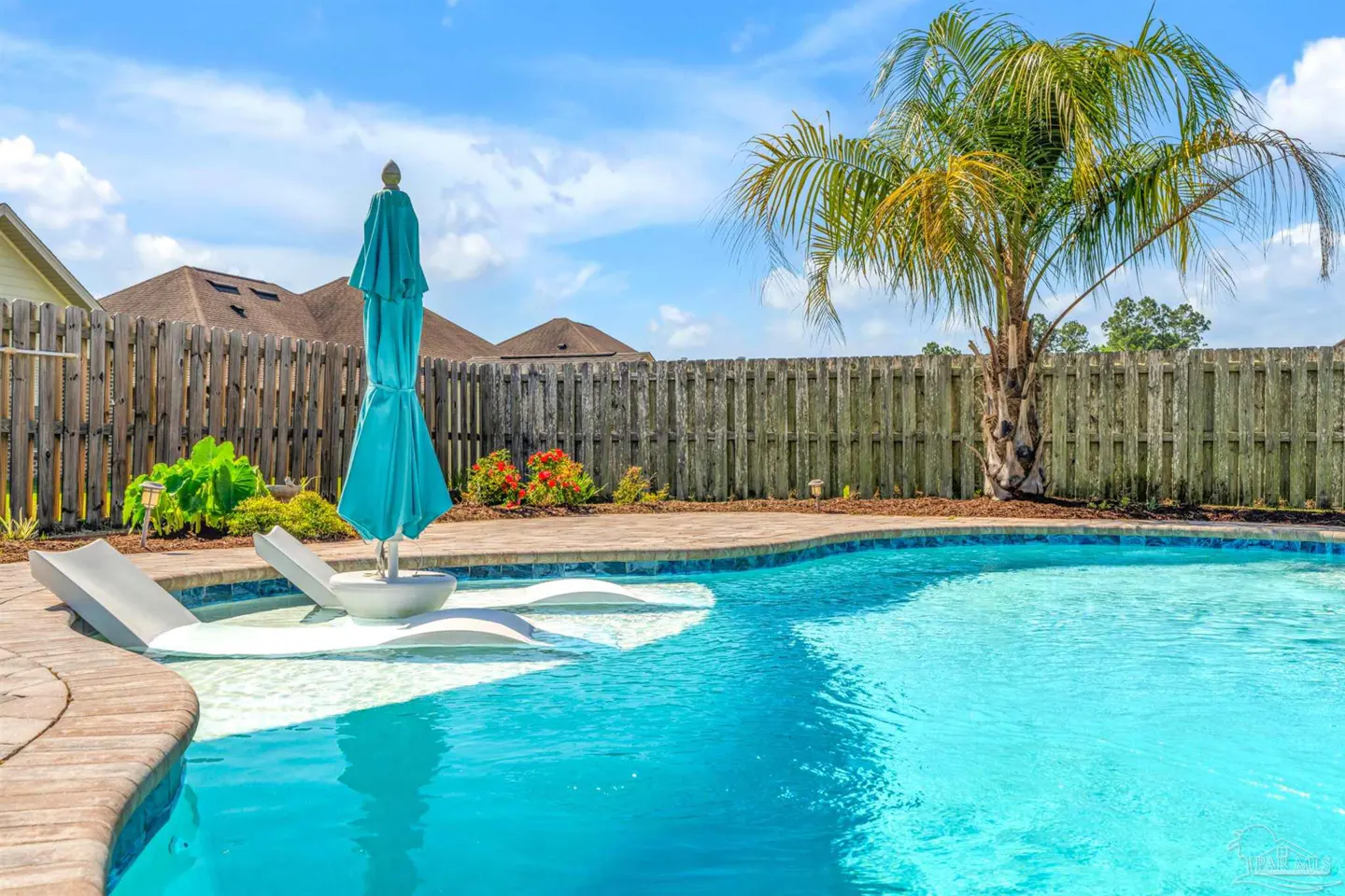 A backyard pool with lounge chairs, a closed turquoise umbrella, and a palm tree against a wooden fence.