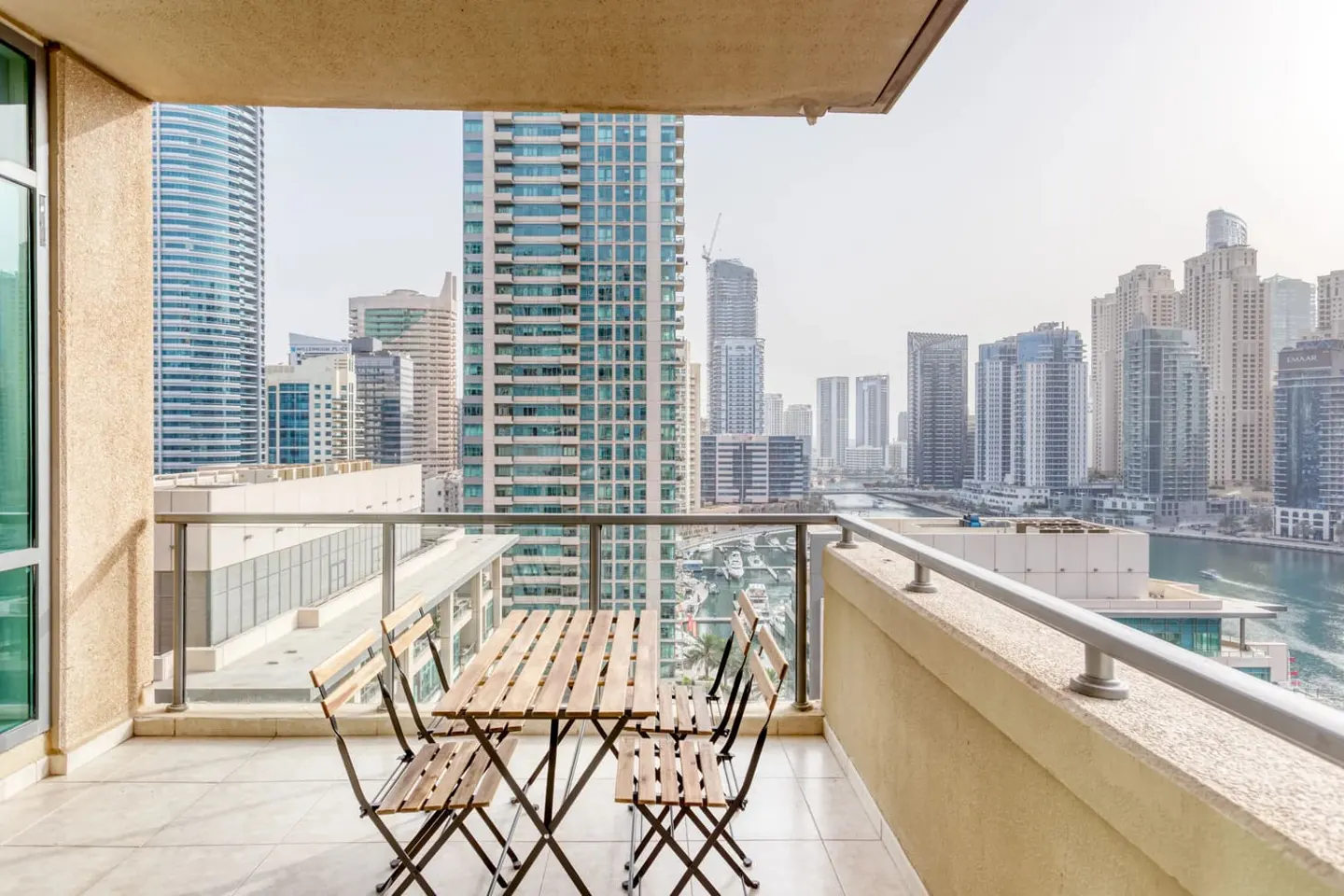Balcony view of Dubai skyline. A wooden table and four chairs sit on a tiled balcony overlooking skyscrapers and a waterway.